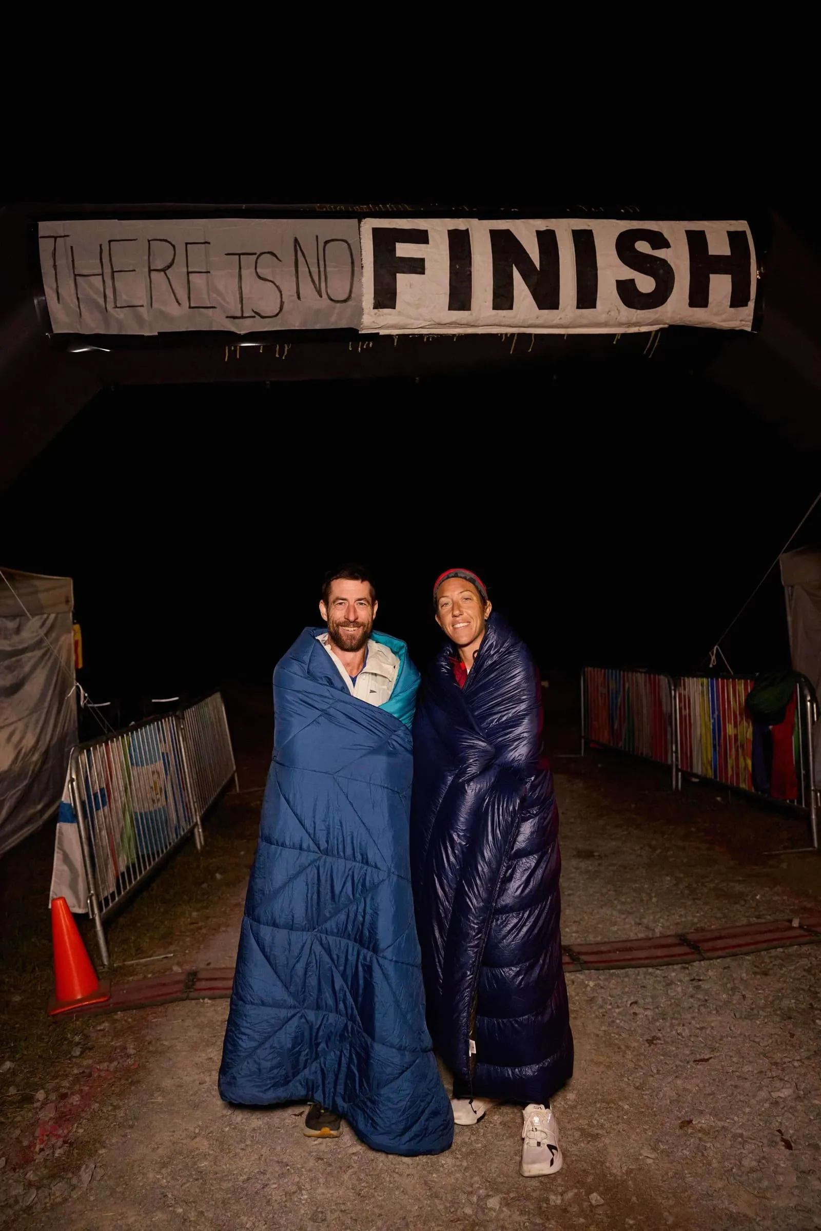Megan Eckert and another runner wrapped in sleeping bags at night beneath a banner reading There Is No Finish