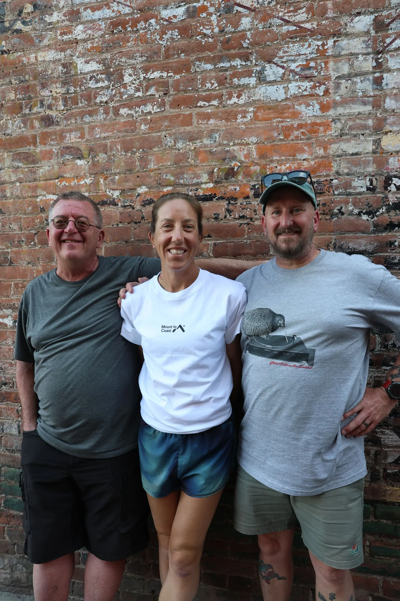 Megan Eckert smiling between her husband Joe Eckert and friend Mike Dobies, arms around each other in front of a weathered brick wall