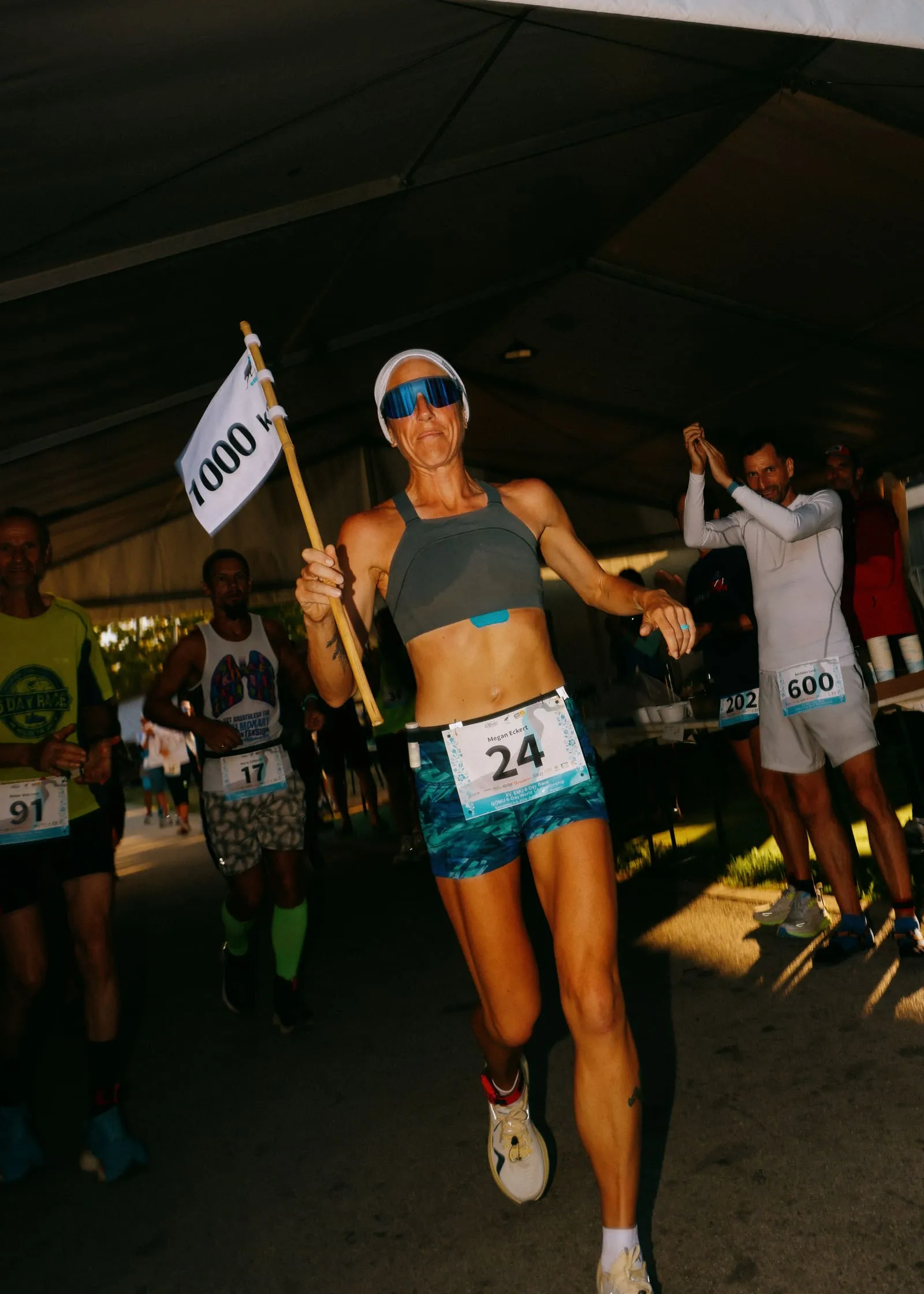 Megan Eckert, bib 24, smiling as she runs carrying a white flag marked 1000 under the race tent at the EMU 6-Day Race in Hungary