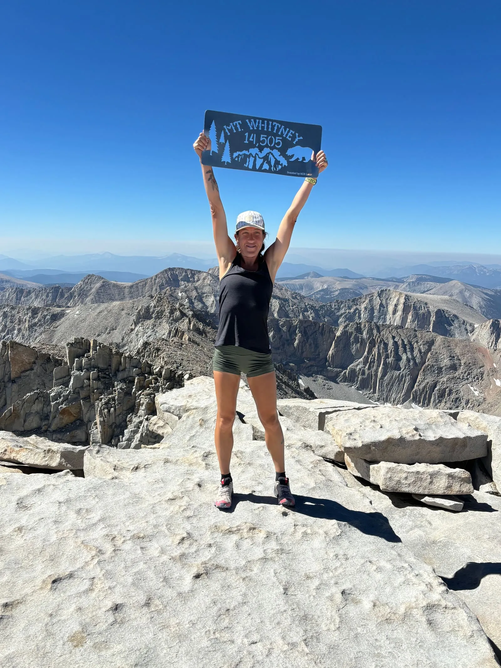 Megan Eckert on the summit of Mount Whitney holding the 14,505 ft sign overhead, with jagged peaks below